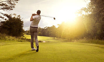 A golfer swings a club on a well-manicured course, surrounded by lush greenery and trees, as the sun sets, casting a golden hue over the scene.