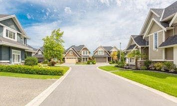 Suburban houses line both sides of a curved street, with manicured lawns and trees in front yards, under a partly cloudy sky.