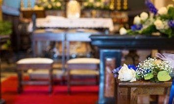 A wooden pew adorned with white and blue flowers sits in a church interior, facing an altar draped with white cloth and surrounded by floral arrangements. Two chairs are aligned before the altar.