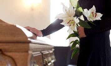 A person holds white lilies and rests a hand on a wooden casket in a well-lit room, suggesting a solemn or memorial setting.