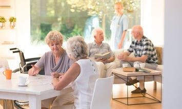 Elderly women sit and interact at a table with a cup and papers; other older adults converse nearby on a couch in a bright, well-lit room with large windows and a healthcare worker.